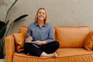 Colleen Goodacre, Registered BAS Agent and Xero Bookkeeper at Cloudscribe, sitting on an orange sofa holding a notebook.